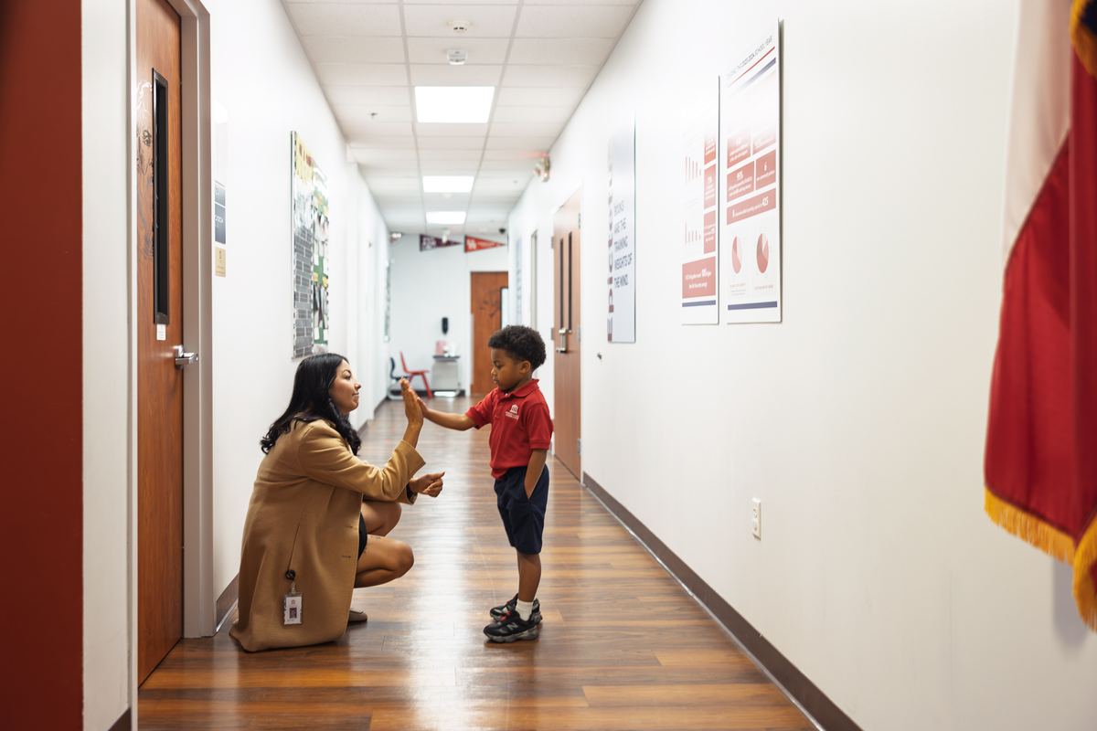 An educator kneeling to give a young student a high five in a school hallway
